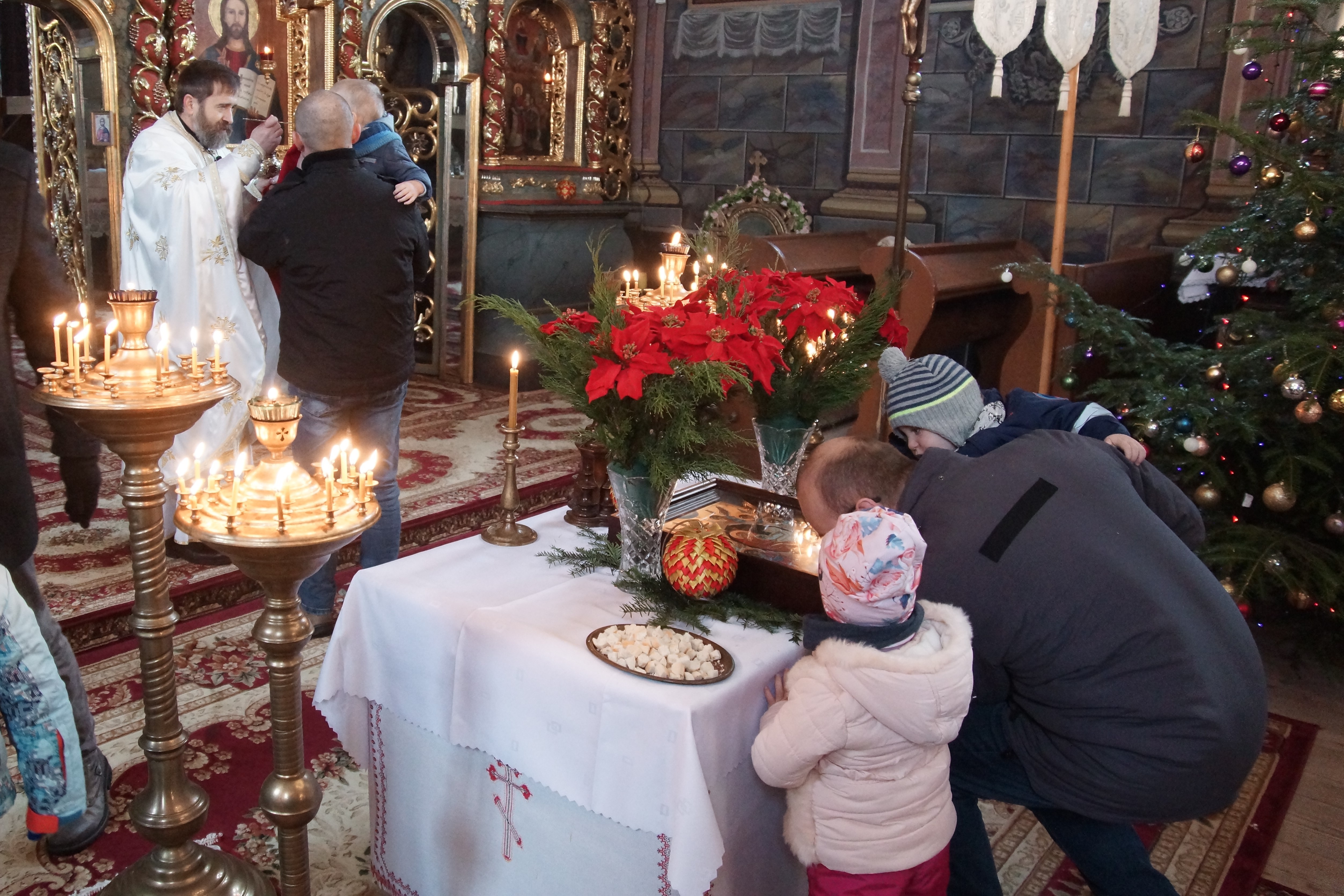 Wnętrze cerkwi. Mężczyzna z dzieckiem na rękach całuje ikonę Bożego Narodzenia leżącą na niewielkim stoliku.